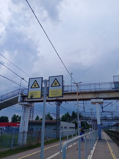 The image depicts a view of a railway station platform, likely in Fryazevo, Russia, under a cloudy sky. Two triangular warning signs are prominently displayed, indicating "Движение скоростных поездов!" (High-speed train traffic!). The signs feature a silhouette of a train within a yellow triangle. A pedestrian overpass, constructed of concrete and metal, crosses overhead, with railings along its sides. The platform itself is paved with interlocking bricks and has a yellow line marking the edge. In the background, there are various railway tracks, electrical wires, and some buildings, including a red and white structure with the partial word "MA" visible, possibly a store or industrial building. Several individuals are present on the platform, some standing and others appearing to walk. The scene is captured from a low angle, looking upwards towards the overpass and the sky.