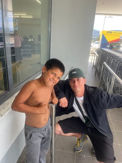 Two males, an adult and a child, are posing for a photo outdoors, seemingly at the entrance of a supermarket. The image is taken at an angle, making it appear rotated.

The adult male, on the right, wears a dark jacket over a white t-shirt with a black stripe, dark shorts, and dark sneakers with yellow laces and soles. He has a dark green baseball cap with "WASCS" (partially visible) written on it and is smiling slightly, looking directly at the camera. He is leaning against a metal railing, which extends into a row of shopping carts behind him.

The child, on the left and slightly in front, is bare-chested and wears grey sweatpants. He is broadly smiling and looking at the camera, also leaning against a lower part of the same or a similar metal railing.

Behind and to the left of the subjects is a large glass storefront or window, reflecting the interior of the store, where fluorescent lights and a faint "LIDL" logo are visible. Another person is reflected inside the store, bending over a shopping cart.

In the background, beyond the shopping carts and the building, an ATM is visible with prominent yellow and blue branding. Text on the ATM reads "ATM" and "ΧΩΡΙΣ ΧΡΕΩΣΗ" (meaning "WITHOUT CHARGE" or "FREE OF CHARGE"). Another Greek phrase, "ΚΑΡΤΕΣ ΠΕΛΑΤΩΝ" ("CUSTOMER CARDS"), is also visible. Beyond the ATM, there are distant hills or mountains under what appears to be a clear sky. The overall impression is a bright, daytime setting. Both individuals appear happy and relaxed.