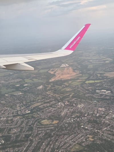 An aerial photograph, likely taken from the window of an airplane, shows a view of St Albans, United Kingdom. The wing of a Wizz Air plane is visible on the left side of the frame. Looking downwards, there is a landscape consisting of green fields, trees, and urban development. Visible is also a large area of bare earth, likely a construction site. In the foreground, there's a dense residential area with houses, streets, and patches of green. The environment appears hazy.
