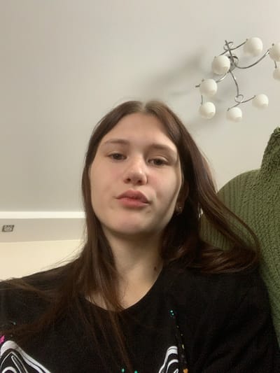 A young woman with long, dark brown hair is captured in a close-up selfie, looking directly at the camera. She is wearing a black t-shirt with a white graphic design and appears to be indoors. The background includes a white ceiling with a modern chandelier and a textured green surface, possibly a chair or sofa. The lighting is soft, suggesting an indoor environment. There are no explicit indicators of the city of Belogorsk, Russia, within the image itself.