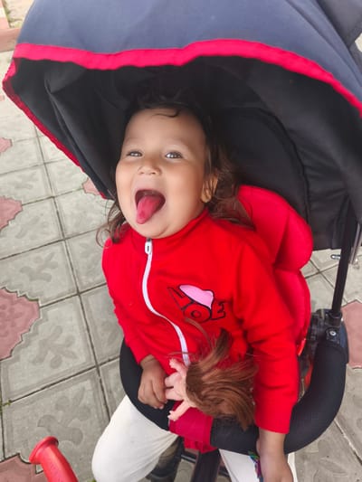 A young child, likely female due to the doll and ear piercing, is seated in a stroller. She is wearing a red hooded jacket and white pants. Her tongue is sticking out, and she is looking towards the camera with wide, dark eyes. Her hair is dark and wavy. A doll with brown hair is held in her right hand. The stroller has a black canopy with a red trim. The background consists of paved ground with a decorative pattern of light and dark gray tiles. The scene appears to be outdoors, possibly in a park or public area. There are no visible landmarks that would specifically place this image in Burayevo, Russia.