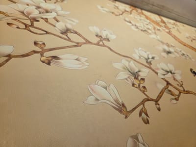 A close-up, angled view of a textured beige fabric displays a detailed illustration of magnolia branches with white and pinkish blossoms. The painting, rendered in a soft, artistic style, features delicate petals and brown branches adorned with buds and open flowers. The fabric has a subtle woven texture that is visible throughout the image, creating a shimmering effect under the light. The composition is dominated by the floral motif, with branches extending diagonally across the frame. The scene does not depict any people or specific urban elements, and therefore, cannot be located within the city of Dĩ An, Vietnam.