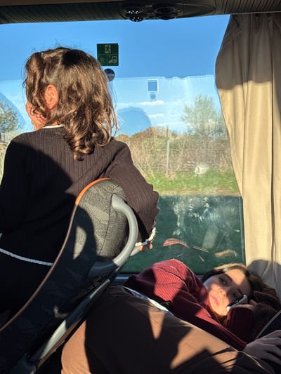 The scene depicts the interior of a passenger bus or coach, located in Ladispoli, Italy, during daylight hours. Two females are present. The first individual, viewed from behind in the foreground, has curly dark brown hair and wears a black ribbed long-sleeved top with white trim on the cuffs and hem. She is seated, looking out the window towards the right. The second individual is positioned in the mid-ground, lying prone across a seat, facing the viewer. She wears a maroon ribbed long-sleeved top and brown pants, and holds a white smartphone with her left hand near her face. Bright sunlight illuminates the interior, casting prominent shadows from the dark grey patterned seats, which feature orange piping and grey plastic handles. An overhead panel with circular vents and lights is visible. Through the large window, a clear blue sky, green and brown vegetation, and a fence are discernible in the background. Safety pictograms, including a green 'exit' sign, are affixed to the window.