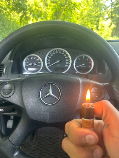 The image shows a close-up view of a car's steering wheel and dashboard. A person's hand is holding a lighter that is lit, with the flame illuminating a button on the steering wheel. The steering wheel has a Mercedes-Benz logo in the center and various buttons for vehicle controls. The dashboard features a speedometer and other gauges, with visible text including "AIRBAG" and "R". The background suggests an outdoor setting with green foliage, implying it is daytime. The overall scene is captured from inside the car, looking towards the steering wheel and dashboard. The specific location is identified as Konstancin-Jeziorna, Poland. The activity is the use of a lighter near the steering wheel controls.