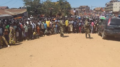 A large crowd of people gathers in a street, bordered by buildings. Two uniformed soldiers stand guard, directing the crowd or providing security, near a black SUV. The setting appears to be a town or city in a developing country, with a mix of informal street-level commerce and more established buildings in the background. The atmosphere appears tense.