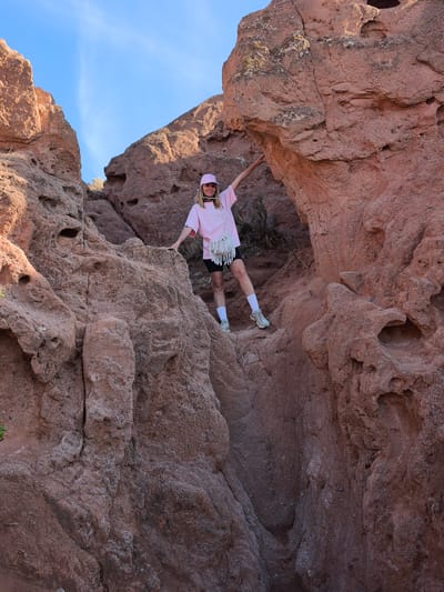 A medium shot captures a fair-skinned person, identified as female, with shoulder-length blonde hair, wearing a pink baseball cap, light pink T-shirt, black athletic shorts, white socks, and grey sneakers. She carries a white fringed shoulder bag. She is positioned between two large, rough, reddish-brown rock formations, holding onto them with one hand each. The background features a clear, light blue sky. The location is Tosor, Kyrgyzstan.