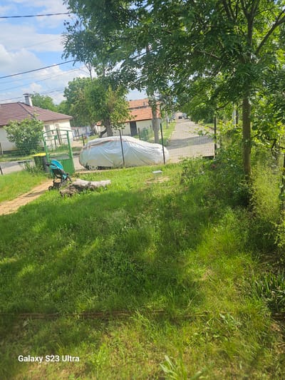 An overhead residential scene in Yana, Bulgaria, captured during daytime under a partly cloudy blue sky. The foreground consists of a vibrant green grassy area, with a faint horizontal metal rail visible towards the bottom. Moving towards the midground, a light brown dirt path leads from the left, past a light blue baby stroller, to a green metal gate in a chain-link fence. A yellow-lidded trash bin is positioned near the gate. To the right of the path and within the fenced area, a car is parked, fully covered by a light grey tarp. Logs or wood pieces lie on the grass near the path. The background reveals a paved street, likely asphalt, with multiple parked vehicles further down. Houses with brown tiled roofs are visible on both sides of the street, including a light-colored house with a chimney on the left and another building on the right. Numerous trees with dense green foliage are prominent throughout the image, particularly a large tree trunk on the right side of the fence and smaller trees near the houses. Utility lines traverse the upper portion of the scene against the sky. No individuals are present.