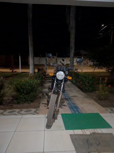 A motorcycle is parked on a tiled walkway at night in front of a building with palm trees and vegetation. The motorcycle is a dark color, and its headlight is on. The walkway is bordered by a low wall and a patch of grass with red flowers. In the background, there is a building with a fence and some overhead power lines. The scene is illuminated by artificial light, likely from the building or streetlights. The location is specified as Cáceres, Brazil.
