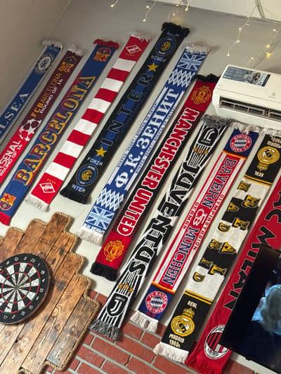This media file features a collection of football scarves, likely belonging to a fan or collector, displayed in an indoor setting. The scarves represent various prominent European football clubs, including AC Milan, Real Madrid, Bayern Munich, Juventus, Manchester United, Inter Milan, FC Zenit, FC Barcelona, Arsenal, and Chelsea. They are arranged in a layered fashion, cascading downwards, with some hanging from what appears to be a wall or a stand.
The background includes elements of a domestic setting, with a white air conditioning unit prominently visible in the upper left corner. Decorative fairy lights are also present, strung along the top edge of the image, suggesting a cozy or festive atmosphere. In the lower right quadrant, a dartboard is mounted on a rustic wooden plank surface, adding a recreational element to the scene. The overall impression is one of personal fandom and a collection curated with care, likely within a home or a dedicated fan space. The lighting suggests an indoor environment, possibly during the day or with artificial lighting. The arrangement of the scarves and the presence of the dartboard hint at a casual, yet organized display of sports memorabilia.