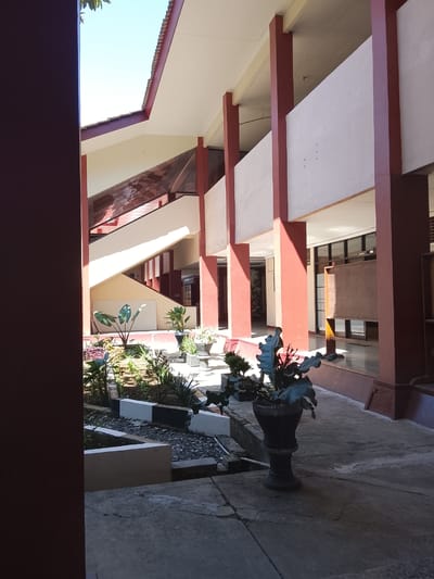 The image shows an outdoor walkway and garden area of what appears to be an academic institution in Karangwangkal, Indonesia. The architecture features a covered walkway supported by red pillars, with cream-colored walls and a white ceiling. A staircase with red railings and cream-colored steps is visible in the background, leading to an upper level. Sunlight casts shadows on the concrete walkway, highlighting a small garden bed with various plants and decorative stones. Several potted plants are placed along the walkway, including a large, dark green leafy plant in an ornate black pot. In the distance, there are windows and doors, suggesting indoor spaces. The overall scene depicts a well-maintained outdoor area conducive to pedestrian traffic and relaxation.