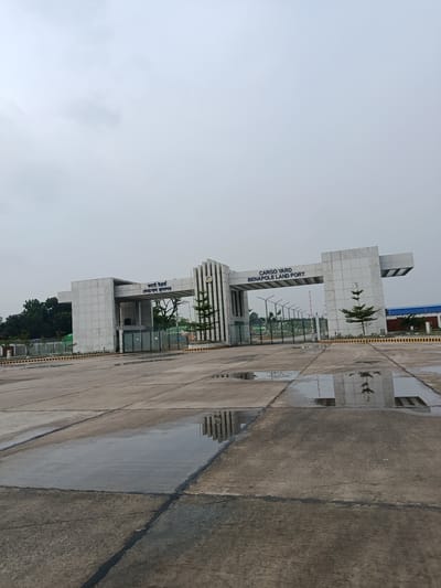 The image shows the entrance to the Benapole Land Port Cargo Yard in Benapole, Bangladesh. The entrance is a large, modern structure with concrete pillars and a horizontal beam. Blue text on the beam reads "CARGO YARD BENAPOLE LAND PORT" in English and Bengali. A green metal gate is visible behind the entrance, with a security checkpoint beyond it. The ground in front of the entrance is a paved concrete area with several puddles of water reflecting the sky and the entrance structure. Trees and foliage are visible on the left side of the image, and a blue building is partially visible on the right. The sky is overcast with clouds. There are no people visible in the image. The scene appears to be a functional port facility under a cloudy sky.