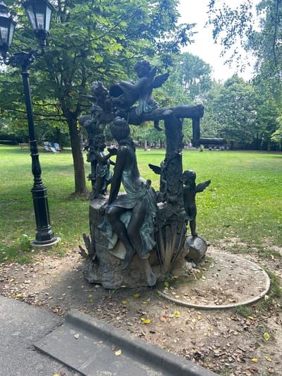 The image depicts a bronze sculpture of cherubic figures and a woman in a park setting. A dark, ornate lamppost stands to the left of the sculpture, which is surrounded by a grassy area with scattered leaves. In the background, more trees are visible, along with lounge chairs and a fence. The scene is likely outdoors in a public park, given the presence of the sculpture, lamppost, and manicured lawn.