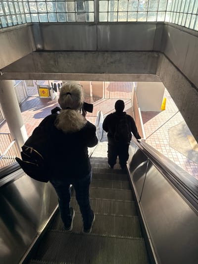 A downward-angle photograph depicts two individuals descending an escalator at a Metrorail station in Hialeah, United States of America. In the immediate foreground, an adult female with light-colored hair, styled in a ponytail, wears a dark jacket, blue jeans, and a black backpack with visible gold zippers. She holds a dark electronic device, possibly a camera or smartphone, to her right eye, aimed forward. Ahead of her, a second, silhouetted adult individual, dressed in dark clothing and carrying a backpack, occupies a lower step on the escalator. The escalator features ribbed metal steps and a stainless steel handrail. Above, the station's ceiling structure consists of concrete beams and translucent square glass blocks, allowing diffused natural light. The brightly lit lower level reveals a patterned reddish-brown tiled floor, white metal railings, a yellow caution cone positioned on the right side, and a yellow 'No Entry' sign with a red circle and white bar, affixed to a barrier on the left.