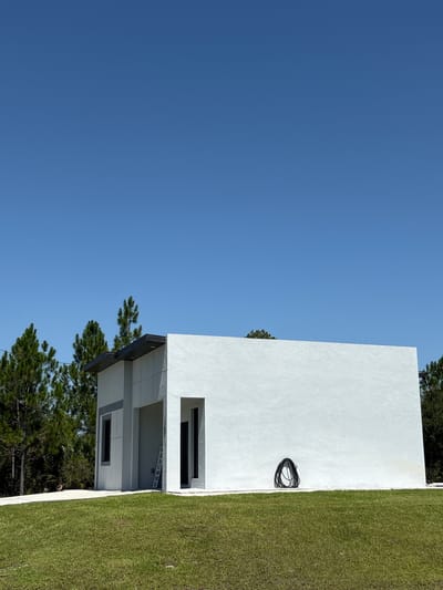 A modern, minimalist white stucco building stands in a grassy field under a clear blue sky in Lehigh Acres, Florida, USA. The building features sharp angles and recessed dark windows. A black coiled hose rests against the right side of the structure. Pine trees border the background, and a ladder is visible next to the building's entrance. A concrete path leads up to the building.