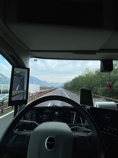 The image depicts the interior of a Volvo truck cab, providing a view of the road ahead through the windshield. The highway is bordered by barriers, and beyond the road, mountains can be seen. The speedometer indicates a speed of 82 km/h. A Pepsi-themed air freshener is suspended from the windshield. A screen displays a view from a side-view camera. The photo was likely taken in Nave San Rocco, Italy, based on location information.