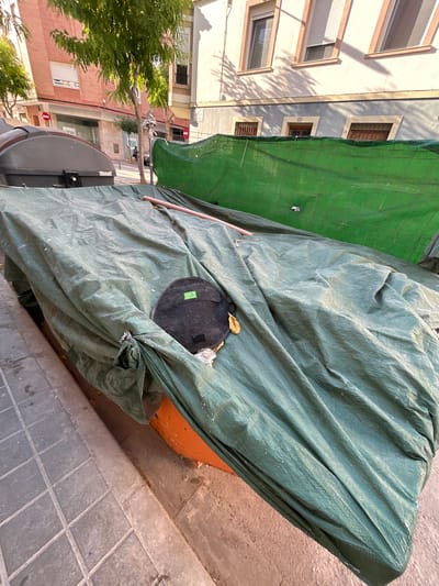 This is an eye-level, full shot of a street scene in Castelló De La Plana, Spain, during the day. The image is dominated by a large, green tarp covering what appears to be an outdoor object or structure. The tarp is heavily wrinkled and draped, suggesting it is being used to protect something from the elements or for storage. A thin, light-colored pole is visible poking out from under the tarp.
On the tarp, a circular, dark-colored object with a green tag that reads "TOP" is placed. Next to it, a small, dry, yellow leaf rests on the green material. The tarp itself is draped over an orange-colored surface, hinting at the object it is covering.
To the left of the tarp, a portion of a building is visible. It has a light-colored facade with several windows, some of which have wooden shutters. Sunlight casts distinct shadows of tree branches onto the building's walls, indicating it is a sunny day. Further to the left, the leaves of a lush green tree are visible.
The ground in the foreground is paved with square, grey tiles. The scene appears to be on a sidewalk or street. The overall impression is one of an urban outdoor space, possibly undergoing some form of construction or renovation, or simply storing items. The lighting suggests it is midday. There are no people visible in the frame.