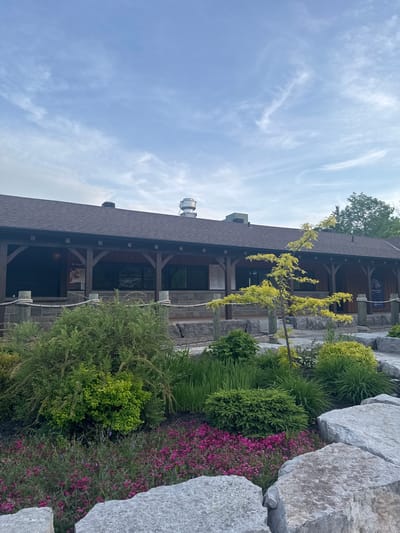 The image, correctly oriented, presents an outdoor scene featuring a rustic-style building surrounded by natural landscaping under a bright, partly cloudy sky. The main subject is a long, single-story building with a brown shingled roof and dark wooden support beams and pillars, suggesting a lodge, visitor center, or similar establishment. A white vent or chimney is visible on the roof towards the left.

In the foreground and mid-ground, a meticulously designed landscape features large, light gray boulders prominently on the right side. Various green plants and shrubs, including dense evergreens, tall grasses, and bushy deciduous plants, are arranged around these rocks and along the front of the building. Patches of vibrant pink and purple flowering plants add color to the garden beds. A small tree with striking bright yellow-green foliage stands in front of the building, near the center. Rope barriers are visible along the front of the building, indicating a pathway or designated area.

The sky is a clear light blue with scattered, wispy white clouds, suggesting a pleasant daytime setting. No people are present, and no specific activity is taking place.

Visible text on signs attached to the building includes a partially visible blue sign with a white logo that appears to say "discovery [something]". Another sign below it has "2023 C[...]" and other small print. Further left, a sign contains the text "EB College Str" or similar, and possibly the number "6175". The scene evokes a tranquil, natural environment, characteristic of areas around Chelsea, Canada.