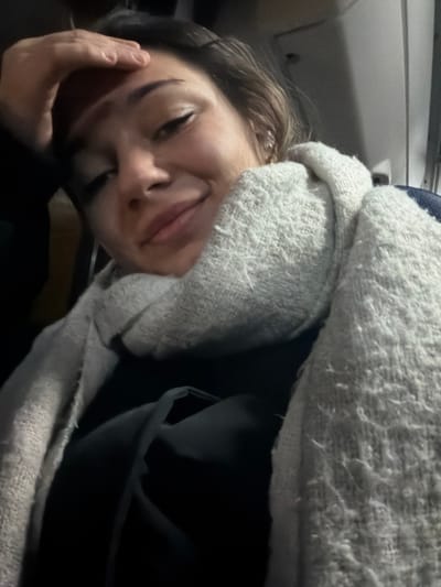 A person's face, eyes closed, is partially obscured by a fuzzy white blanket. The individual's head rests on their hand, and the background suggests they might be in a vehicle, possibly a car or plane. The blanket is wrapped around the person, indicating comfort or sleep. The lighting is subdued, adding to the sense of rest.