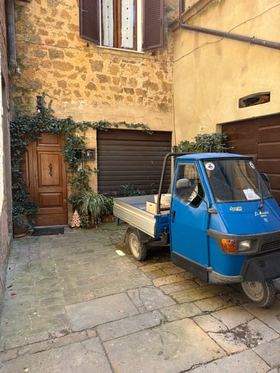 This media file depicts a scene in Pienza, Italy, likely during the daytime with bright sunlight casting shadows. The image features a bright blue three-wheeled Piaggio Ape 50 utility vehicle parked on a cobblestone street. The vehicle has a cargo bed in the back. The setting is an old building with a textured stone facade and a wooden door with ornate carvings. Green ivy vines creep up the side of the building, partially obscuring the door and the vehicle. A window with decorative white fabric coverings is visible on the stone wall. Another window with a brown slatted cover is present above the stone wall.

Text visible on the vehicle includes "Ape 50" and "La Motoria", which appears to be a brand or company name. There are also stickers on the vehicle's windshield. A small plaque with the number "58" is affixed to the building near the ivy. The overall impression is a charming, rustic street scene in a historic Italian town. No people are visible in the image.