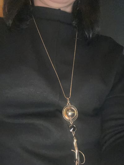 A gold necklace with a circular pendant and a small black gemstone is draped over a black garment, possibly a coat or dress. The necklace's chain is thin and delicate. The garment's texture is smooth and dark, providing a stark contrast to the jewelry. Some fur or feathers are visible on the left side of the image, also attached to the garment.
