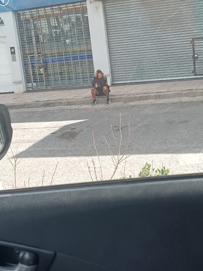 A seated person with their head in their hands sits on a curb between a building with a barred window and a closed roll-up door. They are dressed in dark shorts and a long-sleeved dark shirt. The person is facing away from the viewer and appears to be distressed. The photograph is taken from inside a car with the door panel visible at the bottom. The scene is viewed through a dry, weed-filled patch.