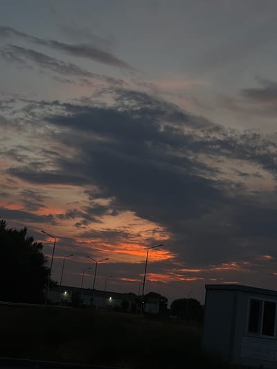 The image captures a dramatic sunset over Aidemir, Bulgaria. Dark, heavy clouds dominate the sky, with streaks of vibrant orange and pink peeking through, indicating the sun is low on the horizon. In the foreground, silhouetted trees line the left side of the frame. A row of streetlights stands tall, their lamps unlit, leading towards a long, low building with illuminated windows in the middle ground. To the right, a small, white, prefabricated structure is visible, with text on its side. The ground in the foreground appears to be covered in grass and vegetation, with a paved area in the lower left corner. The overall atmosphere is twilight, with the last remnants of daylight giving way to darkness.