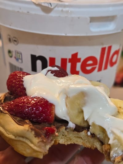 The image displays a close-up, slightly rotated view of a delectable dessert. The main subject is a section of a golden-brown waffle or thick pancake, generously topped with fresh, vibrant red strawberries and slices of ripe yellow banana. A substantial dollop of fluffy white cream, likely whipped cream or yogurt, cascades over the fruit and the dessert base. A hand is partially visible on the right, holding the sweet treat.

In the blurred background, a large white container of Nutella is prominent, with its iconic red "Nutella" logo clearly readable. Below the logo, some smaller, indistinct icons are also visible on the container. The cross-section of the dessert reveals its soft texture and hints of dark chocolate spread, presumably Nutella, layered within. The overall impression is one of a rich, indulgent, and appetizing dessert. This sweet scene takes place in La Florida, Chile.