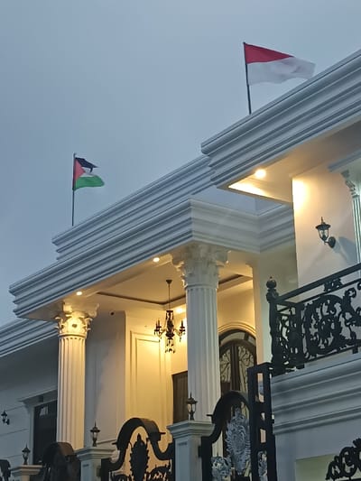 This image depicts a section of a large, ornate building situated in Sindangasih, Indonesia, under twilight or nocturnal conditions. The structure features white or light-colored facades with classical architectural elements, including fluted columns topped with Corinthian capitals, supporting a decorative entablature and roofline.

Two national flags are displayed on flagpoles positioned on the roof. On the left, the Palestinian flag, consisting of black, white, and green horizontal stripes with a red triangle at the hoist, is visible. On the right, the Indonesian flag, with red over white horizontal stripes, is present. Both flags are partially unfurled.

The building's entrance area is illuminated by multiple artificial light sources. Warm-toned recessed lights are embedded within the ceiling of the portico, and several dark-colored, decorative wall sconces are mounted on the facade. A multi-armed, dark-toned chandelier with dangling elements hangs from the center of the portico ceiling. A black security camera is mounted on the portico ceiling above one of the columns.

In the foreground, an ornate black metal gate or fence, characterized by intricate scrollwork, partially obscures the lower portion of the building. A decorative black metal balustrade is visible on the right side of the image, suggesting an upper-level balcony or terrace. The sky above is a uniform light gray, indicative of dusk or an overcast evening. No individuals are visible in the scene.