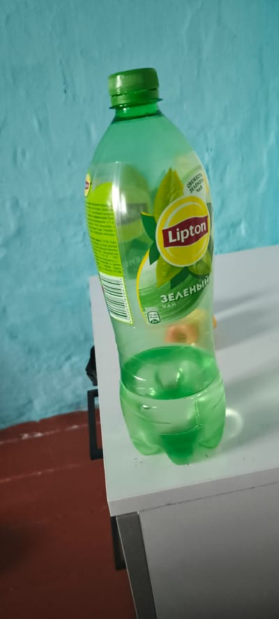 A green plastic bottle of Lipton green tea is positioned on a white surface. The bottle is partially filled with a light green liquid. The bottle has a green cap and features a label with the Lipton logo, which is a red "Lipton" text inside a yellow oval surrounded by green leaves. Text in Russian, including "ЗЕЛЁНЫЙ ЧАЙ" (green tea), is also visible on the label. The background consists of a textured light blue wall and a dark reddish-brown floor. The scene appears to be indoors, likely in a home or office setting, based on the furniture and wall. The bottle is the primary subject of the image.