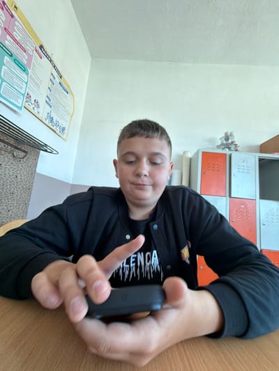 A young boy is sitting at a wooden desk in a classroom, holding a dark-colored electronic device. He is wearing a black jacket over a white t-shirt with an image on it. The background shows a wall with educational posters, lockers, and some rolled-up papers. The classroom appears to be in Zmeitsa, Bulgaria, possibly within a school setting due to the presence of lockers and posters. The boy's focus is on the device in his hands.