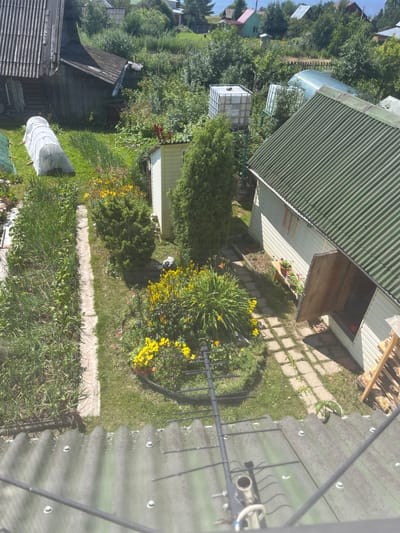 The image shows a rural yard in Gari, Russia. In the foreground, there is a corrugated metal roof with an antenna. The midground features a garden with various plants, including flowers and vegetables, a small white shed with a green roof, and a larger building with a green roof. In the background, there are more houses and trees, suggesting a residential area. The scene appears to be set on a sunny day. There are no people visible in the image.
