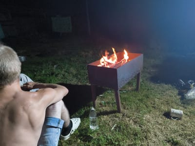 The image shows a person sitting on grass next to a barbecue grill with a fire burning in it. The scene is outdoors at night, with some smoke or fog present. The person is shirtless and wearing denim shorts and white clogs. There is a plastic bottle and a mug on the ground near the grill. In the background, there is a dark area that could be a building or trees. There is also a light-colored object hanging in the background. The overall impression is of a casual gathering or activity taking place outdoors in the evening. The specific location is stated as Патоцкі сельскі Савет, Belarus, which suggests a rural or semi-rural setting.