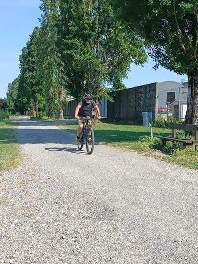 An adult male is observed riding a dark-framed mountain bicycle on a light-colored gravel path in Pieve Saliceto, Italy. He is wearing a dark short-sleeved shirt, light-colored shorts, a black cycling helmet, sunglasses, and light-colored shoes. His hands are on the handlebars, and his feet are positioned on the pedals. The sun, located to the right, casts distinct shadows of the cyclist and his bicycle onto the gravel surface to their left.
The gravel path extends into the background, veering slightly to the left. It is bordered on both sides by green grass and low vegetation. To the left of the path, a continuous line of tall trees with green foliage frames the scene. To the right of the path, a tall, dark, weathered concrete barrier or wall runs parallel to the path. Behind this wall, a light gray industrial-style building with a curved roof and associated fencing and gates are visible.
Within the grassy strip between the path and the wall on the right, a gray cylindrical waste receptacle mounted on a post is present, along with a dark wooden park bench with a backrest. The sky above is clear and blue.