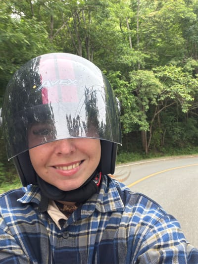 In Samoeng Tai, Thailand, a person wearing a black and pink helmet with a reflective visor is captured in a selfie. The visor reflects trees and the sky, obscuring the upper part of their face. The person is smiling broadly, revealing their teeth. They are wearing a blue and white plaid shirt over a tan garment. Their hair, light brown, is visible on the side, and a black neck protector is worn. The background shows a paved road curving through a lush, green forest with tall trees. The road has a yellow line marking the edge. The image appears to be taken from a moving vehicle, likely a motorcycle, given the perspective and the person's attire.