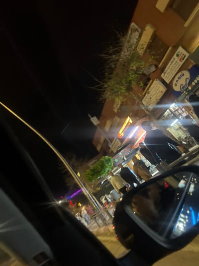 A nighttime photograph taken in Beni Mellal, Morocco, from the interior of a vehicle. The lower left and bottom center show elements of a car's dashboard or door panel, while the lower right features a prominent reflection in the passenger-side mirror. The scene depicts a brightly lit urban street. Multi-story buildings with commercial storefronts, illuminated by artificial lighting, line the right side of the street. Store signs displaying text in Arabic and French, including "Le Foyer d'Oasis," are affixed to the building facades. A prominent metal barrier extends along the sidewalk. Several adult individuals are present on the sidewalk, observed walking or standing in proximity to the storefronts. Scattered trees with sparse to moderate foliage are visible near the buildings and along the street. The sky above is dark, indicating night conditions.