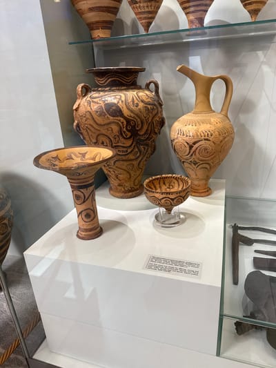 The image displays a museum exhibit featuring ancient Minoan pottery in Heraklion, Greece. In the foreground, on a white pedestal, are several clay vessels. To the left is a tall, flared chalice with intricate dark brown spiral and linear patterns. Next to it is a small, rounded bowl with similar decorative motifs, resting on a clear acrylic stand. Behind these, a large, bulbous amphora dominates the center, adorned with dark brown curvilinear designs resembling octopus tentacles and swirling patterns. To the right of the amphora stands a tall, narrow-necked jug with a handle, also decorated with concentric circles and wave-like designs. The pottery rests on white display surfaces within a glass case. In the background, on a higher shelf, more pottery with banded and geometric patterns is visible. To the right of the display case, part of another exhibit featuring dark metal tools or weapons can be seen. A descriptive label is visible on the white pedestal, providing information about the artifacts. The environment appears to be an indoor museum setting with neutral-colored walls and overhead lighting. No people are present in the frame.