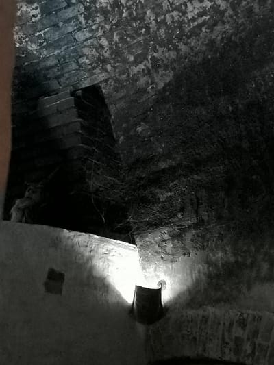The image depicts the interior of the Kuldīga Old Town, specifically within the Kuldīga Castle. The scene is dimly lit, with the primary light source appearing to be a lamp or flashlight casting a strong, focused beam onto a rough-hewn wall. The architecture suggests an aged and possibly underground structure, with visible brickwork on the ceiling and upper walls, exhibiting signs of wear and discoloration. There is a distinct sense of depth and shadow, creating an atmospheric and somewhat foreboding environment.
In the foreground, a rough, light-colored wall runs horizontally across the lower portion of the image. To the right of the wall, a dark, cylindrical object, possibly a bucket or a container, is illuminated by the light source, which is positioned nearby and directed upwards. The beam of light highlights the texture of the wall and casts sharp shadows.
Further back and to the left, a darker area reveals more of the brick structure. A stepped, rectangular feature made of dark bricks is visible, partially obscured by shadow and possibly cobwebs. The overall impression is one of an old, possibly disused or storage area within the historic Kuldīga Castle. The image is devoid of any people or explicit human activity, focusing solely on the architectural elements and the play of light and shadow.