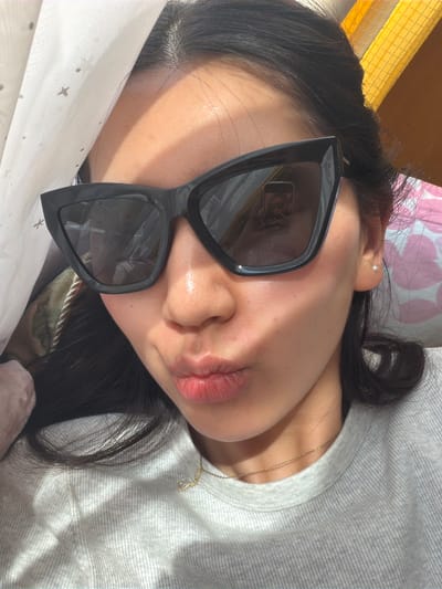 A close-up, angled shot features a person lying down, facing upwards with their head tilted slightly to the right. The person is wearing large, dark-framed sunglasses that dominate the view of their face. A gold chain necklace is visible on their neck. They have dark hair and a light complexion. The background includes a white surface with small patterns and a gray, textured fabric. There are also hints of a yellow object and a pink floral-patterned fabric in the periphery. There is nothing in this image to suggest a location in the city of Kawasaki, Japan.