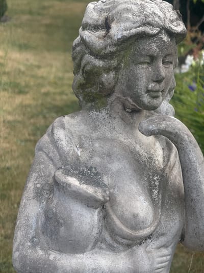 A weathered, grey stone statue of a woman is positioned outdoors, against a blurred backdrop of green grass and foliage. The statue depicts the woman from the chest up, with her head slightly tilted to her right and her right arm bent, with her hand resting on her chin. Her hair is styled in an ornate updo. The stone shows signs of age, with patches of moss and discoloration, suggesting it has been exposed to the elements for a significant period. The lighting suggests it is daytime. The statue is located in Heidelbach, Germany.
