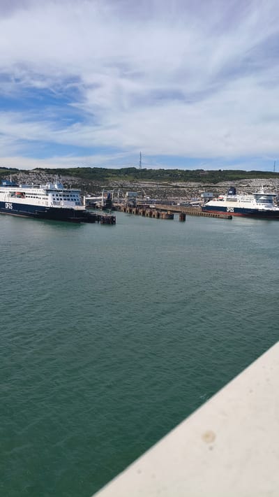 This image captures a view of the Dover Ferry Port in Dover, United Kingdom, on a clear day with a partly cloudy sky. Two large ferries, identified by the "DFDS" logo, are docked at the port. The ferry on the left is primarily white with a dark blue hull, and the ferry on the right also features white and dark blue with a red stripe. The ferries are connected to the port infrastructure, which includes several piers and loading ramps. Behind the port, a green, hilly landscape rises, topped with chalk cliffs characteristic of the White Cliffs of Dover. A tall communications tower stands on the hill. The sea in the foreground is a deep teal color, with small ripples on its surface. A portion of a white structure, possibly the railing of the viewpoint from which the photo was taken, occupies the bottom right corner of the frame. There are no people clearly visible in the image.