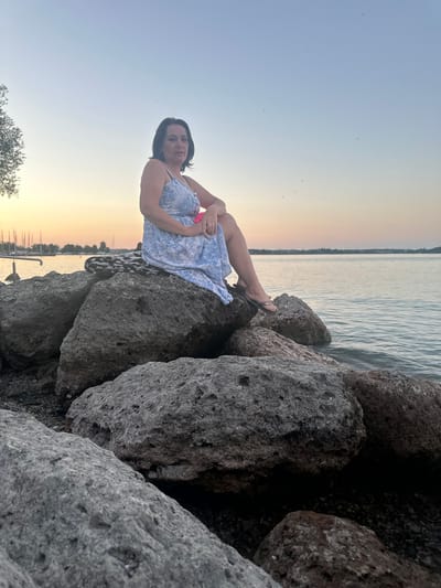A middle-aged woman with dark hair and a blue and white patterned dress is sitting on large rocks by a lake. She is looking towards the camera with a neutral expression. The sky is a soft gradient of blue and orange, indicating either sunrise or sunset. The lake is calm and reflects the colors of the sky. In the background, there are sailboats docked and trees lining the shore. The foreground is dominated by the rough texture of the rocks. The scene appears to be located in Prien am Chiemsee, Germany, given the presence of a lake and sailboats, common features of this Bavarian town.