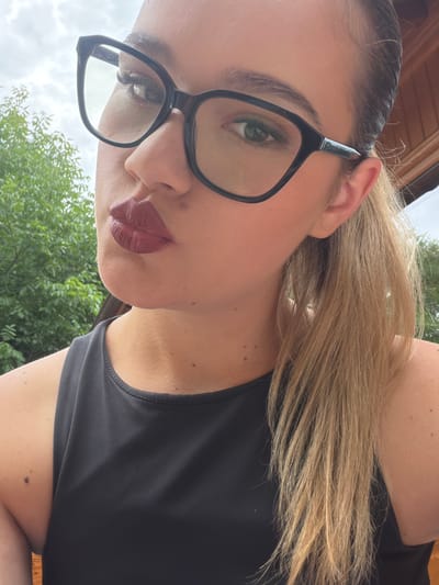 This media file is a close-up, self-portrait of a woman. She is wearing large, black-rimmed glasses and has dark red lipstick on her lips. Her blonde hair is pulled back into a ponytail. She is wearing a black tank top. The image is taken outdoors, with greenery and what appears to be a cloudy sky in the background. There is also a wooden structure visible at the top of the image, suggesting an outdoor setting like a patio or deck. The lighting suggests it is daytime. No text is visible in the image. The photograph appears to have been taken from a low angle looking up at the subject.