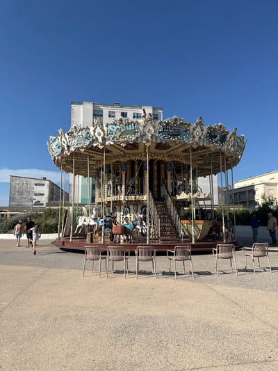 A carousel with painted horses and decorative elements stands outdoors on a paved area under a clear blue sky. The carousel is gilded and has a stepped entrance. In the foreground, several empty chairs are arranged in a row. In the background, there are buildings, some of which appear to be apartments, and greenery. A few people are walking on the paved area to the left of the carousel. The scene is likely located in the city of Berck, France, a coastal resort town known for its beaches and Belle Époque architecture.