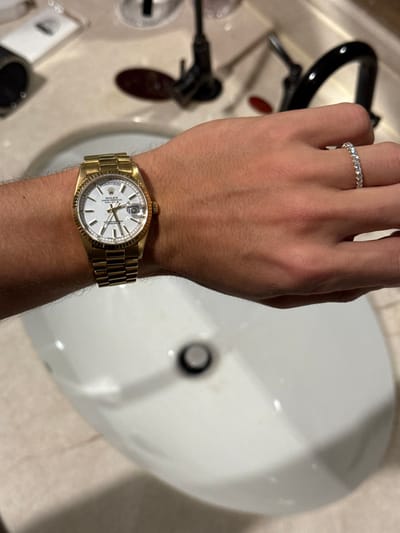 A close-up, high-angle image captures a person's left forearm and hand positioned over a white bathroom sink. The individual wears a gold-toned Rolex Oyster Perpetual Datejust wristwatch on the left wrist, featuring a white dial, fluted bezel, and a gold Jubilee bracelet. The date window on the watch displays the numeral "10". A silver-toned or white gold eternity band ring, set with numerous clear stones, adorns the fourth finger of the left hand. The white ceramic sink, containing a dark circular drain stopper, occupies the lower and central portion of the frame. In the blurred background, a dark-colored, single-handle faucet is visible behind the sink, situated on a light-toned countertop. Additional indistinct objects, including what appears to be a grooming tool, are present in the upper left background. The specific location within the city of Tongxiang, China, is not discernible from the image content.