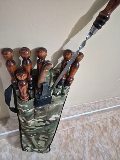The image shows a camouflage bag filled with several wooden-handled skewers and a knife. The bag is standing upright against a light beige wall, with a tiled floor in the foreground. The skewers have twisted metal ends, and the knife is sheathed in a black holder. The lighting suggests an indoor environment. The image does not provide any information about the location in Tskhinvali, South Ossetia.