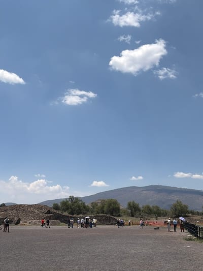 The image shows a partially obscured view of the archeological site of Teotihuacan in San Juan Teotihuacán, Mexico. A long stone structure, presumed to be part of the ancient city, runs from the bottom center to the top right, flanked by a wide, gravel-like pathway. Numerous people, likely tourists, are scattered along the pathway and near the structure, engaging in walking and observation. The environment also includes trees and a distant, somewhat hazy mountain range. The sky is a clear blue, dotted with scattered white clouds.