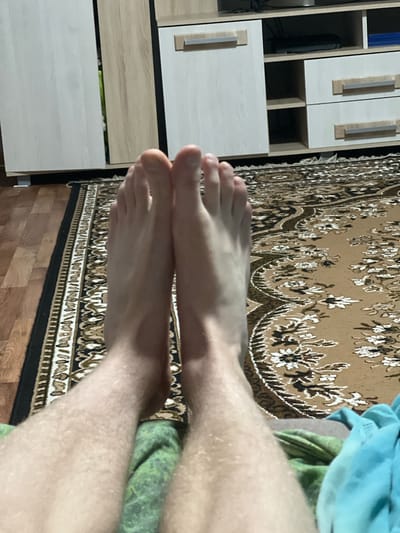This is a close-up, eye-level shot of a pair of bare feet and lower legs positioned on a rug. The feet are facing upwards, with toes spread slightly. The lower legs are covered in light-colored hair. A green cloth is visible between the legs, and a blue cloth is partially visible to the right. In the background, there is a wooden floor and a piece of furniture with white and light wood cabinets, suggesting an indoor domestic setting. There is no indication that this image is located in Gorshki, Russia.
