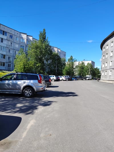 A street scene in the city of Olenegorsk, Russia, features a parking lot filled with various vehicles and bordered by apartment buildings and trees under a clear blue sky. The foreground shows the asphalt surface of the parking lot with shadows cast by trees and a silver SUV parked on the left. Several other cars, including a black SUV, a white compact car, a blue sedan, a dark pickup truck, and a white van, are parked further back in the lot. To the left, a long, multi-story apartment building with numerous windows stretches across the frame, partially obscured by green trees. To the right, a curved, multi-story building with light-colored brickwork and white-framed windows is visible. Power lines are suspended in the sky. The scene appears to be a typical residential area in a Russian city.