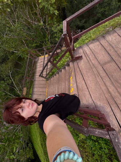 A vertically oriented photograph captures a person on a wooden staircase descending into a dense jungle environment near Flores, Guatemala. The foreground features a light-skinned individual with reddish-brown, medium-length hair, wearing a black t-shirt with a white and red graphic design, and a light blue coiled wristband on their right forearm. This individual's right arm is extended, holding the camera to capture a selfie-style view, with their gaze directed towards the lens. The immediate scene shows the upper section of a robust wooden staircase, constructed with weathered brown planks and dark brown wooden railings. Faded yellow paint marks are visible along the edges of several stair treads and landings. The staircase recedes downwards, surrounded by thick tropical foliage consisting of various green trees and undergrowth. A small, rectangular wooden sign with "SIEMPRE CON SU PECHO" inscribed in dark lettering is visible on the left side railing at a lower segment of the stairs. A faint, distant human figure can be discerned further down the descending staircase.