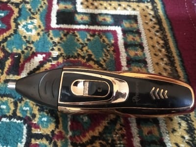 The scene is observed in Asaka, Uzbekistan. A black and gold "ProGamer" electric nose and ear hair trimmer is positioned horizontally on a multi-colored patterned rug. The trimmer features a silver-colored cylindrical cutting head at its left end. The main body is black plastic with gold-colored metallic accents, including a gold-framed slider switch located centrally on the upper surface. The switch appears to be in a neutral or off position. The right side of the trimmer displays the "ProGamer" logo and three gold chevron symbols. The rug exhibits a traditional, intricate pattern composed of teal, maroon, beige, black, and white elements. No individuals or active interactions are present.